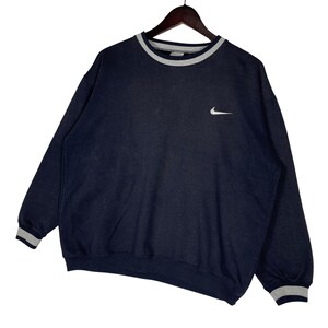 May include: Navy blue sweatshirt with a white Nike logo on the chest. The crew neck, cuffs, and hem feature gray and white striped detailing. The sweatshirt is hanging on a wooden hanger.