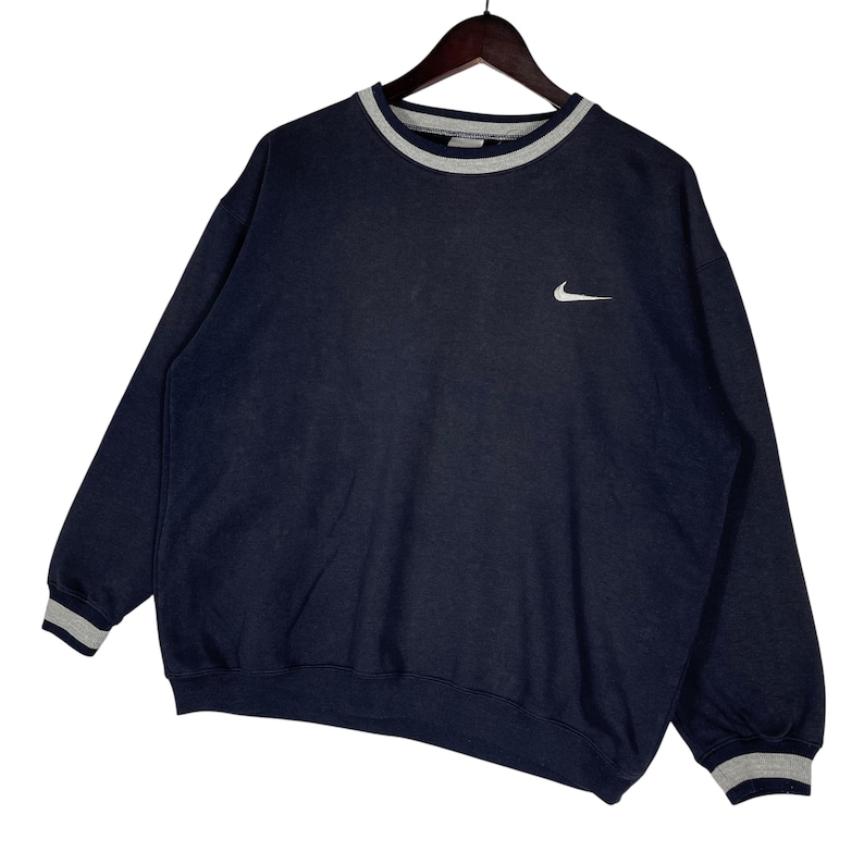 May include: Navy blue sweatshirt with a white Nike logo on the chest. The crew neck, cuffs, and hem feature gray and white striped detailing. The sweatshirt is hanging on a wooden hanger.