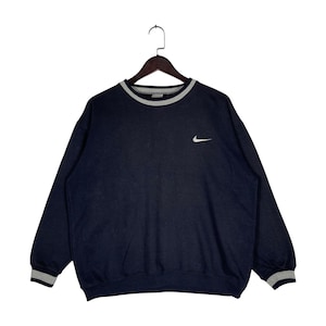 May include: Navy blue sweatshirt with a white Nike logo on the chest. The crew neck and cuffs have a gray and white striped trim. The sweatshirt is hanging on a wooden hanger.