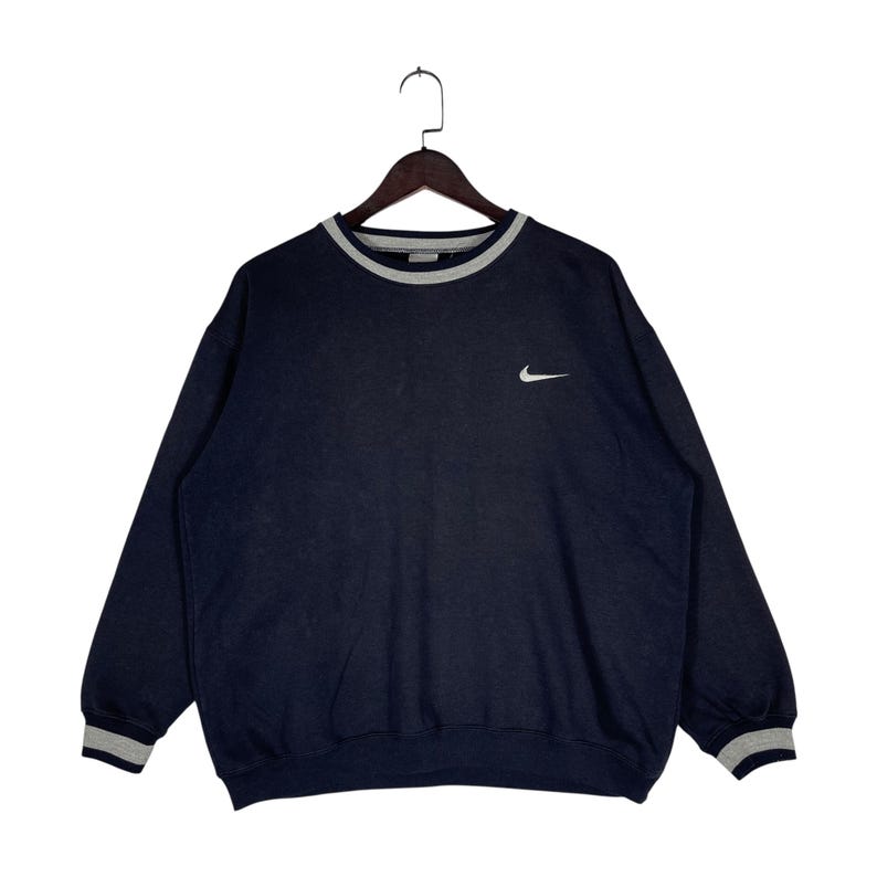 May include: Navy blue sweatshirt with a white Nike logo on the chest. The crew neck and cuffs have a gray and white striped trim. The sweatshirt is hanging on a wooden hanger.