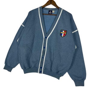 May include: A blue cardigan sweater with white trim and blue buttons. The sweater has a V-neck and two front pockets. A patch with a shield design is on the chest. The sweater is hanging on a wooden hanger.