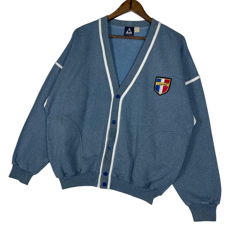 May include: A blue cardigan sweater with white trim and blue buttons. The sweater has a V-neck and two front pockets. A patch with a shield design is on the chest. The sweater is hanging on a wooden hanger.