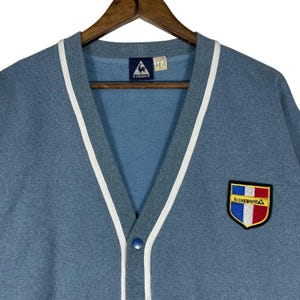 May include: A light blue cardigan with white trim along the edges and a button closure. The cardigan features a patch with the text "le coq sportif" and a rooster logo. The cardigan is hanging on a wooden hanger.