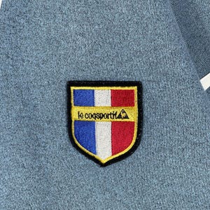 May include: Close-up of a light blue fabric with a colorful patch. The patch is a shield shape with a gold border, featuring a white, blue, and red design, and the text "le coq sportif".
