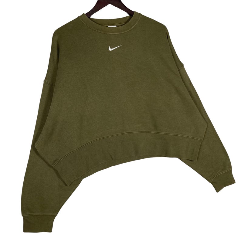 Op de afbeelding: Olijfgroene sweatshirt met een ronde hals en lange mouwen. Voorzien van een wit Nike-logo op de linkerbovenborst. De sweatshirt heeft een unieke, gebogen zoom en hangt aan een houten kleerhanger.