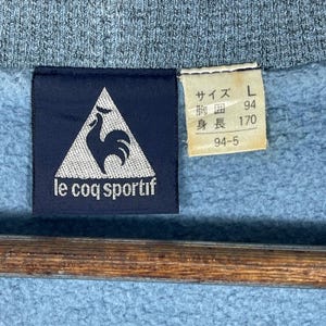May include: Close-up of a blue sweatshirt with a navy blue label featuring the Le Coq Sportif logo, a white rooster inside a white triangle. A smaller tag displays Japanese text and the size L.