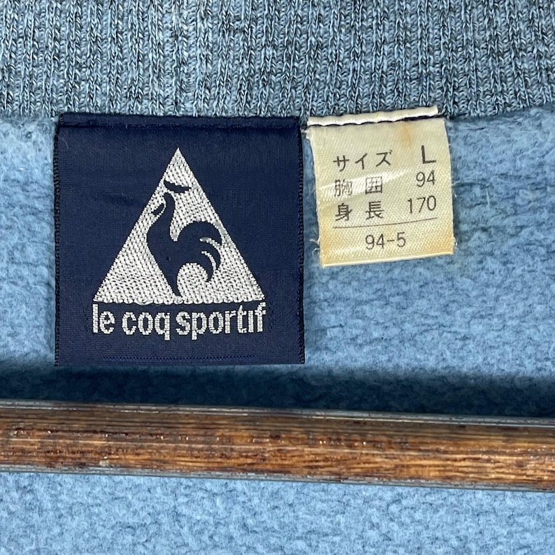 May include: Close-up of a blue sweatshirt with a navy blue label featuring the Le Coq Sportif logo, a white rooster inside a white triangle. A smaller tag displays Japanese text and the size L.