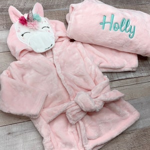 May include: Pink hooded bathrobe with a unicorn applique on the hood. The robe has a tie belt and the name "Holly" is embroidered on the matching robe.