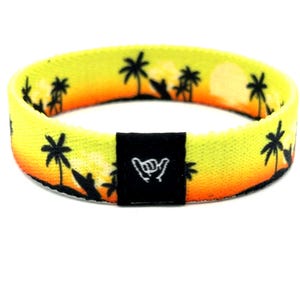 Op de afbeelding: Een stoffen polsbandje met een geel en oranje zonsondergangverloop als achtergrond, voorzien van zwarte silhouetten van palmbomen en surfplanken. Een zwarte rechthoekige stoffen patch is in het midden geplaatst met een wit geborduurd shaka-handgebaar.