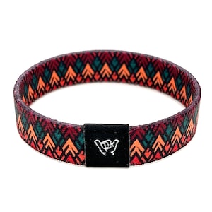Puede incluir: Una pulsera tejida de color rojo, naranja y verde con un cierre ajustable negro que presenta un símbolo de mano "shaka" blanco.
