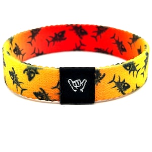 May include: A fabric wristband featuring a yellow-to-orange gradient background with a repeating black fish skeleton pattern. A black rectangular fabric tag is centered on the front, displaying a white embroidered shaka hand gesture icon.