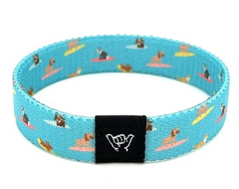 May include: A turquoise elastic bracelet featuring a repeating pattern of dogs surfing on colorful surfboards. A black tag with a white hand gesture is attached to the bracelet. The bracelet is designed to be worn on the wrist.