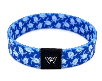 May include: Blue and white patterned wristband with a black adjustable buckle. The pattern features white sea turtles on a blue background. The buckle has a white "shaka" hand sign.