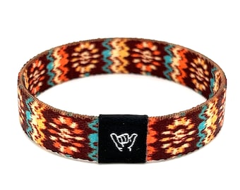 Puede incluir: Una pulsera tejida colorida con un patrón marrón, naranja, amarillo y turquesa. La pulsera tiene un cierre ajustable negro con un símbolo de mano "shaka" blanco.