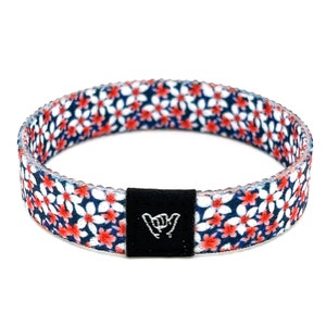 May include: A blue and white patterned wristband with red flowers and a black adjustable buckle with a white "shaka" hand sign.