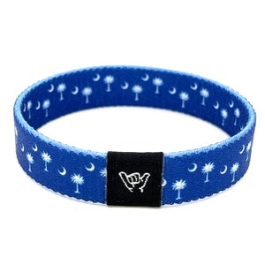 May include: Blue and white woven bracelet with a black adjustable buckle. The bracelet features a repeating pattern of palm trees and crescent moons. The buckle has a white "shaka" hand gesture.