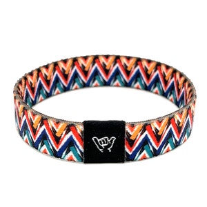 May include: A colorful woven bracelet with a zig-zag pattern in shades of red, orange, blue, and green. The bracelet has a black adjustable buckle with a white "shaka" hand symbol.