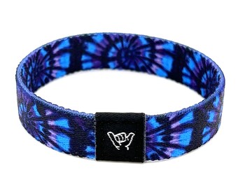 Puede incluir: Una pulsera de tela azul y morada con estampado tie-dye y una hebilla negra ajustable con un gesto de mano "shaka" blanco.