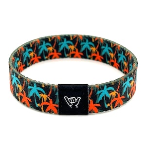 Op de afbeelding: Een zwarte geweven armband met een kleurrijk palmboompatroon en een zwarte verstelbare gesp met een wit "shaka" handgebaar.