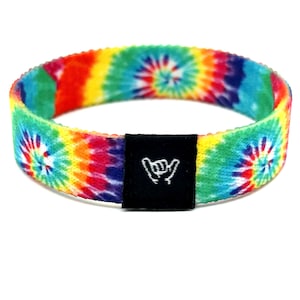 Può includere: Un braccialetto in tessuto circolare caratterizzato da un vivace motivo tie-dye multicolore con vortici di rosso, giallo, verde e blu. Una piccola etichetta rettangolare in tessuto nero è fissata sul davanti, con un segno della mano shaka ricamato in bianco.