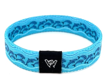May include: A blue and white woven wristband with a pattern of dolphins swimming in waves. The wristband has a black adjustable buckle with a white "shaka" hand gesture.