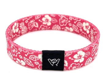 May include: Pink woven bracelet with a white floral pattern and a black adjustable buckle with a white "shaka" hand gesture symbol.