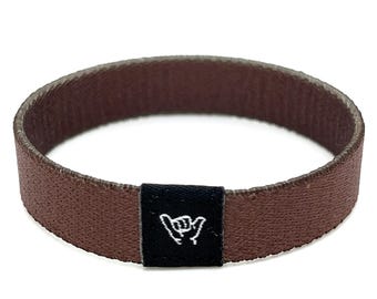 May include: A brown woven bracelet with a black fabric patch featuring a white "shaka" hand gesture.