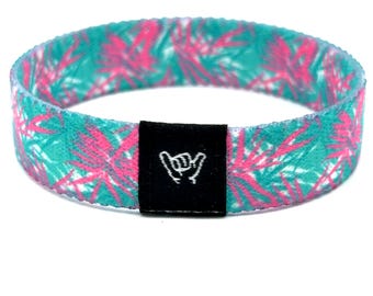 Puede incluir: Una pulsera de tela circular con un patrón de hojas de palma en tonos turquesa vibrante y rosa intenso. Una pequeña etiqueta rectangular negra está cosida en la parte frontal, mostrando un icono gráfico blanco de una mano haciendo un gesto shaka.