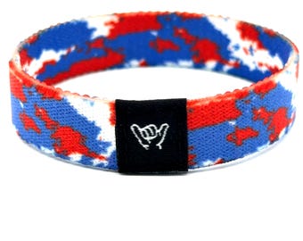Può includere: Un braccialetto elastico in tessuto di forma circolare con un motivo tie-dye rosso, bianco e blu. Un'etichetta rettangolare in tessuto nero è fissata sul davanti, mostrando un'icona bianca ricamata di un gesto della mano shaka su sfondo nero.