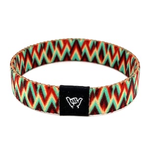 Puede incluir: Una pulsera tejida colorida con un patrón de chevron en tonos de rojo, naranja, verde y marrón. La pulsera tiene un cierre ajustable negro con un símbolo de mano "shaka" blanco.