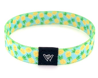 May include: A blue and yellow pineapple patterned adjustable bracelet with a black adjustable buckle featuring a white "shaka" hand sign.