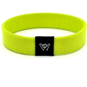 Pode incluir: Uma pulseira elástica amarelo neon com uma pequena etiqueta retangular de tecido preto centralizada na frente. A etiqueta preta exibe um gráfico bordado em branco de uma mão fazendo o sinal de shaka, com dedos curvados e polegar estendido.