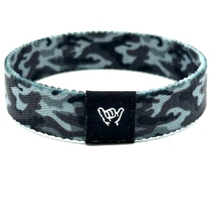 May include: A circular elastic wristband featuring a dark gray and light blue camouflage pattern. A small, rectangular black fabric tag is stitched onto the front, displaying a white embroidered shaka hand gesture icon centered on the band.