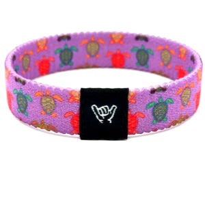 May include: A purple elastic wristband featuring a repeating pattern of small green and orange sea turtles. A black rectangular fabric tag is attached to the front, displaying a white line-art icon of a hand making a shaka gesture.