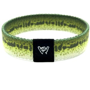 May include: A circular fabric wristband featuring a green and yellow fish scale pattern with a dark green mottled texture. A small black rectangular fabric tag is attached to the front, displaying a white embroidered shaka hand gesture icon.