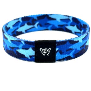 May include: A circular elastic fabric bracelet featuring a repeating pattern of sharks in various shades of blue. A small, rectangular black fabric tag is attached to the front, displaying a white embroidered shaka hand gesture symbol.