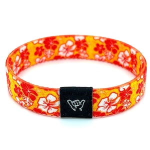 Könnte beinhalten: Ein gelbes und oranges gewebtes Armband mit einem weißen Hibiskusblumenmuster und einem schwarzen verstellbaren Verschluss mit einem weißen "Shaka"-Handzeichen.