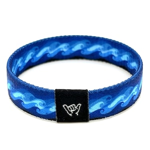 Puede incluir: Pulsera tejida azul y blanca con un patrón de olas y una hebilla ajustable negra con un gesto de mano "shaka" blanco.