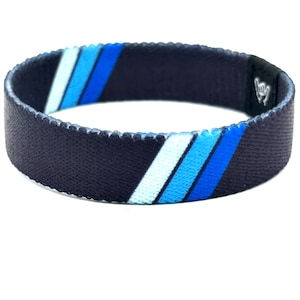 May include: A dark navy blue elastic wristband featuring two diagonal stripes in light blue and medium blue. The band has a textured fabric surface and a small black rectangular patch on the side displaying a white hand gesture icon.