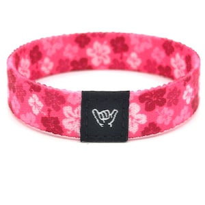 Può includere: Un braccialetto in tessuto rosa con un motivo floreale ripetuto in tonalità di rosa scuro, rosa chiaro e bianco. Una piccola etichetta rettangolare in tessuto nero è cucita sul davanti, mostrando una grafica bianca di un gesto della mano shaka.