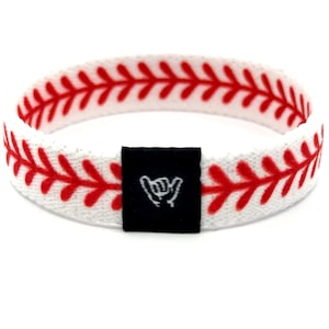 May include: A white fabric wristband featuring a repeating red baseball stitch pattern along the entire length. A small, rectangular black fabric tag is attached to the front, displaying a white line-art icon of a hand making a shaka gesture.