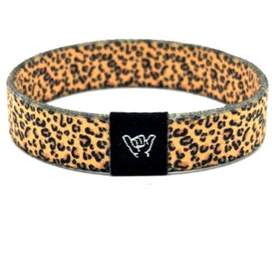 Può includere: Un braccialetto elastico circolare caratterizzato da una stampa leopardata color marrone chiaro e nero. Sulla parte anteriore è applicata una piccola etichetta rettangolare in tessuto nero con un simbolo bianco ricamato a forma di shaka.