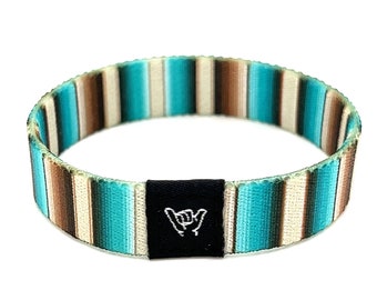 Puede incluir: Una pulsera tejida a rayas turquesa, marrón y blanca con una hebilla ajustable negra que presenta un gesto de mano "shaka" blanco.