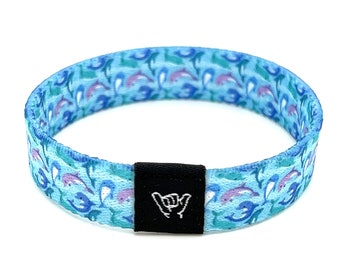May include: A blue and green patterned wristband with a black adjustable clasp. The pattern features white dolphins swimming in a blue and green ocean. The clasp has a white "shaka" hand symbol.