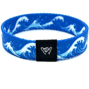 May include: A blue fabric wristband featuring a repeating white ocean wave pattern. The wristband includes a small, rectangular black fabric tag attached to the front, which displays a white embroidered shaka hand gesture symbol.
