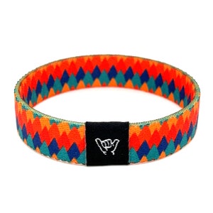 May include: A colorful woven bracelet with a geometric pattern in shades of orange, red, blue, and green. The bracelet has a black tag with a white "shaka" hand gesture.