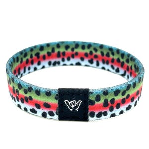 May include: A colorful elastic bracelet with a trout pattern. The bracelet features a design of green, red, and black spots, resembling a trout's scales. A small black tag with a white hand gesture is attached to the bracelet.