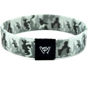 Puede incluir: Una pulsera elástica circular con un patrón de camuflaje en gris, blanco y negro. Una etiqueta rectangular de tela negra está unida al frente, mostrando un icono bordado en blanco de un gesto de mano shaka sobre el material oscuro.