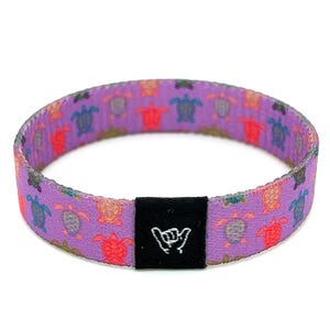 May include: Purple woven bracelet with a black adjustable buckle. The bracelet is decorated with a repeating pattern of colorful sea turtles.