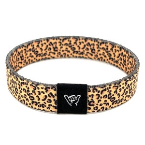 May include: A brown and black leopard print wristband with a black adjustable buckle featuring a white "shaka" hand gesture.